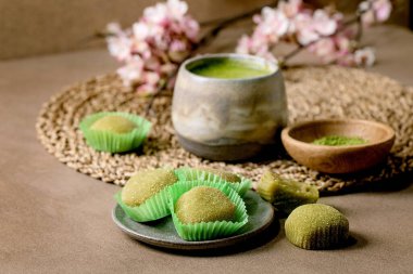 Asya pirinçli tatlı yeşil kibrit mochi, bir fincan köpüklü kibrit çayı ve yeşil kibrit tozu kahverengi masada seramik tabakta. Çaydanlık ve bahar Pembe çiçekli dal arkasında.