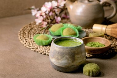 Asya pirinçli tatlı yeşil kibrit mochi, bir fincan köpüklü kibrit çayı ve yeşil kibrit tozu kahverengi masada seramik tabakta. Arkasında çaydanlık ve bahar pembesi açan bir dal var. Boşluğu kopyala