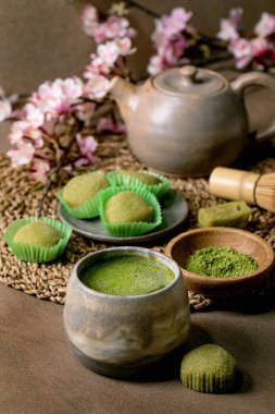 Asya pirinçli tatlı yeşil kibrit mochi, bir fincan köpüklü kibrit çayı ve yeşil kibrit tozu kahverengi masada seramik tabakta. Çaydanlık ve bahar Pembe çiçekli dal arkasında.