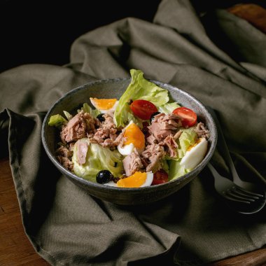 Geleneksel Fransız nicoise salatası, konserve ton balığı, zeytin ve yumurta, seramik kasede, ahşap masadaki keten masa örtüsünde servis ediliyor. Koyu kırsal tarzda. Kare resim