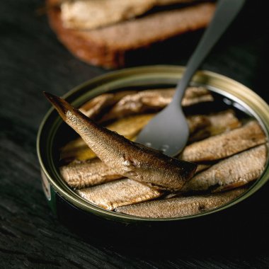 Konserve baltık tütsülenmiş, yağlı, çatallı, arkasında çavdar ekmeği olan siyah ahşap bir masa. Kapatın. Kare resim