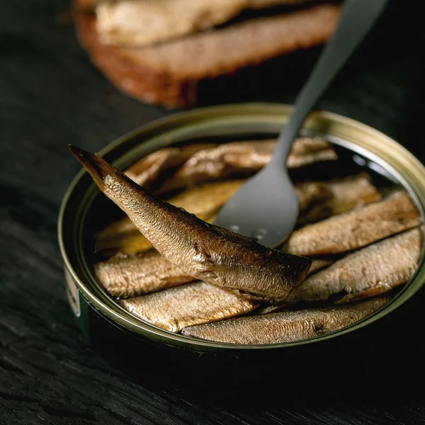 Konserve baltık tütsülenmiş, yağlı, çatallı, arkasında çavdar ekmeği olan siyah ahşap bir masa. Kapatın. Kare resim