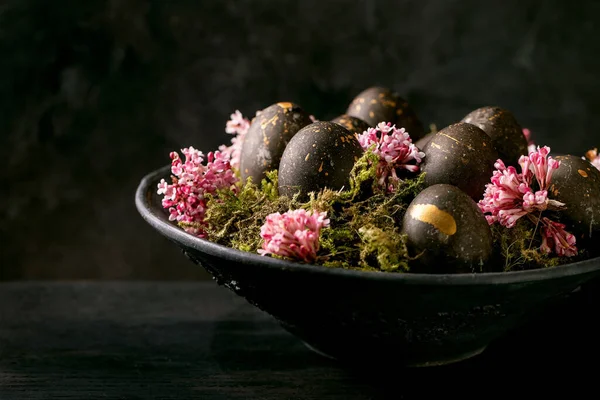 黑色复活节的概念黑色生物彩蛋 金斑点缀在野生苔藓上 粉红的小花插在深色木制背景的黑色陶瓷碗中 — 图库照片