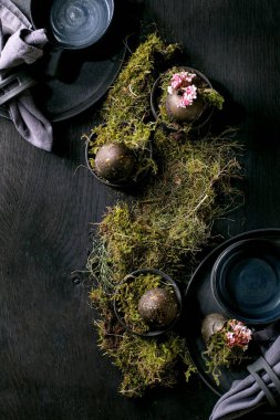 Paskalya sofrası boş seramik tabaklar ve kupalarla süslenmiş, orman yosunlarıyla süslenmiş, siyah paskalya yumurtaları ve ahşap masa üzerinde pembe bahar çiçekleri. Karanlık, şık tatiller. Düz yatıyordu