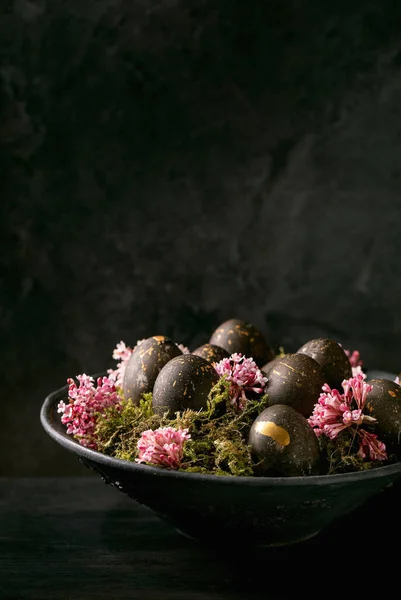 黑色复活节的概念黑色生物彩蛋 金斑点缀在野生苔藓上 粉红的小花插在深色木制背景的黑色陶瓷碗中 复制空间 — 图库照片