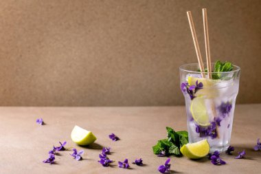 Şeffaf bir bardak limonata soda kokteyli, naneli menekşe çiçekli buz ve limon dilimleri, kağıt pipetler, kahverengi masanın üzerinde duruyor. Soğuk yaz içeceği