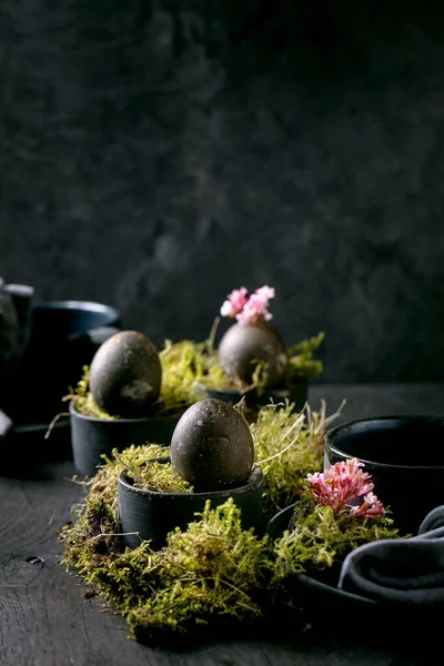 复活节桌上摆满了空的黑色陶瓷盘子和杯子 装饰有苔藓 黑色彩蛋和木制桌子上的粉色春花 黑暗时尚的假期 — 图库照片