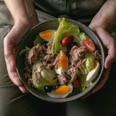 Kadın elinde konserve ton balığı, zeytin ve yumurta ile bir kase nikotin salatası tutuyor. Omega 3 vejetaryen yemeği. Kare resim