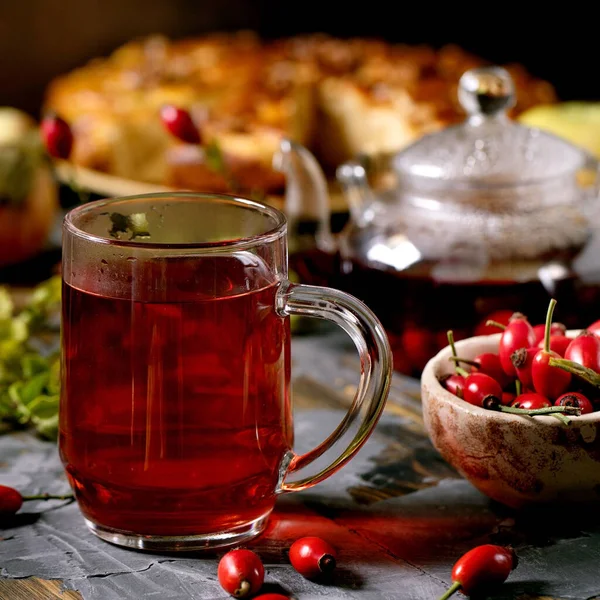 Cam bardakta gül kalçası çayı. Eski ahşap masada duruyor. Yaban güz meyveleri ve elmalı turta da var. Sağlıklı sıcak içecek. Karanlık kırsal bir ruh hali. Kare resim