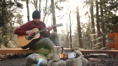 Ormanda ateşin yanında, lacivert şapkalı, gitar çalan genç bir turist.