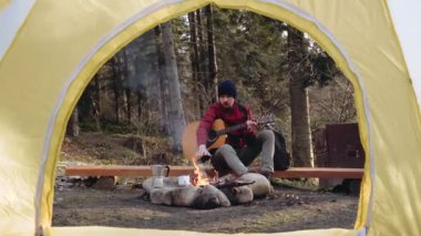 Ormanda ateşin yanında, lacivert şapkalı, gitar çalan genç bir turist.