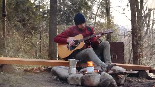 Egy fiatal srác turista az erdőben a tűz mellett, kék sapkában, gitározik. — Stock videók