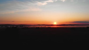 Sakin turuncu bir günbatımında tarlaların sinematik çekimi. Güneş ışınları. Güneşin mavi gökyüzündeki parlak diski yavaşça yükseliyor. Sis