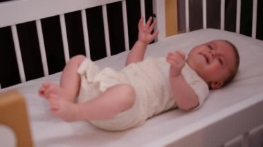 Beyaz tulumlu dört aylık bebek, rahat, ahşap bir karyolada uzanıyor. Bebekler için konforlu modern mobilya reklamı, sağlıklı bebeklik, çocuk bakımı kavramı.