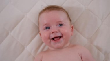 Yeni doğmuş bebek kıyafetleri içinde bebek bezi takmış, çarşafların üzerinde uzanmış ayak parmaklarıyla oynuyor, gülüyor kameraya bakıyor. Kaygısız sağlıklı bebeklik, sağlık ve pediatri konsepti.
