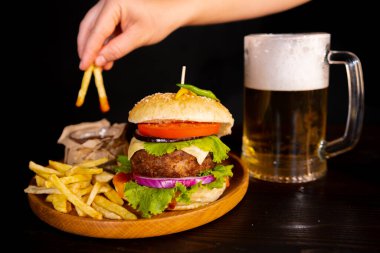 tasty hamburger burger sandwich with french fries and ketchup on a wooden tray a glass of cool beer with foam fast food black background. hand dips french fries in ketchup High quality photo