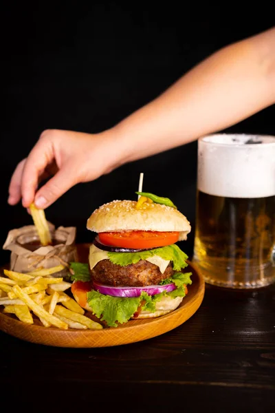 tasty hamburger burger sandwich with french fries and ketchup on a wooden tray a glass of cool beer with foam fast food black background. hand dips french fries in ketchup High quality photo