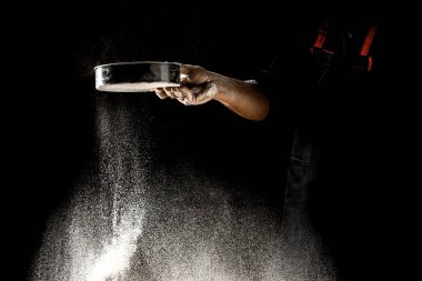 Şef eleğe un serpiyor, toz un havaya karışıyor. Un bulutunun dondurucu hareketinde un taşıyan şefin elleri.