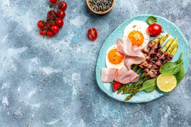 Kahvaltıda yumurta, pastırmaya sarılmış kuşkonmaz, jambon, domates ve salata. Sağlıklı dengeli yiyecekler. Üst görünüm, yukarıda, afiş.