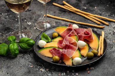 Prosciutto ve fesleğenli taze kavun. Antipasti, geleneksel İspanyolca ve İtalyan aperatifleri. Koyu taş masadaki üst manzara, uzun pankart biçimi. en üstteki görünüm.