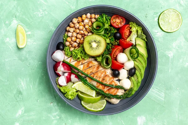 Grilled chicken, avocado, asparagus, chickpeas, broccoli, radish cucumber, tomatoes, olives, mozzarella buddha bowl on green background, top view Delicious balanced food concept.
