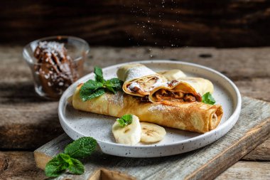 Çikolata kaplı krepler ve beyaz tabakta muz. İnce krep, blini. Lezzetli kahvaltı ya da atıştırmalık. Yemek tarifi geçmişi. Kapat..