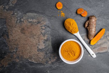 Zerdeçal köklü kaşık içinde zerdeçal tozu. Beton arka planda sokağa çıkma yasağı veya kömür tabakası. Baharat, doğal renklendirme, alternatif tıp. Uzun afiş biçimi. en üstteki görünüm.