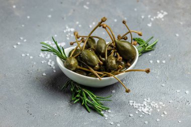 Kapari. Salamura edilmiş ya da turşulu kapari meyvesi. Küçük bir kasede taze biberiye, gri arka planda. metin için yer, üst görünüm.