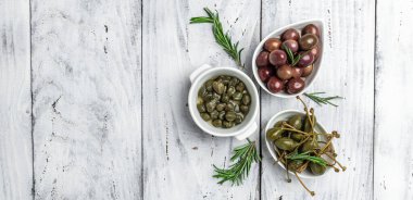 Terbiye edilmiş yeşil ve Kalamata zeytinleri ve kapari, hafif ahşap arka planda, küçük bir kasede taze biberiye. Uzun afiş biçimi. en üstteki görünüm,