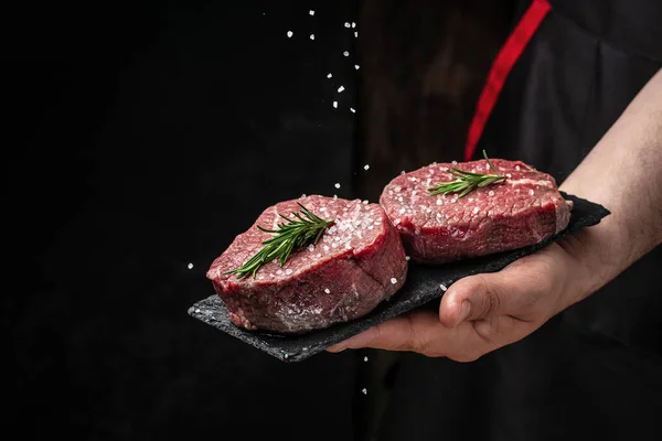 Chef salts steak in a freeze motion with rosemary and spices. Preparing fresh beef or pork on a dark background. Long banner format.