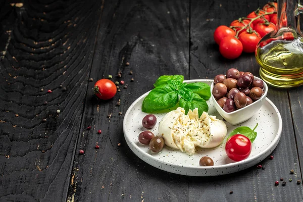 Kremalı İtalyan Burrata peyniri fesleğenli, zeytinyağlı ve beyaz tabakta vişneli domatesli koyu ahşap arka planda. afiş, menü, metin için tarif yeri, üst görünüm.
