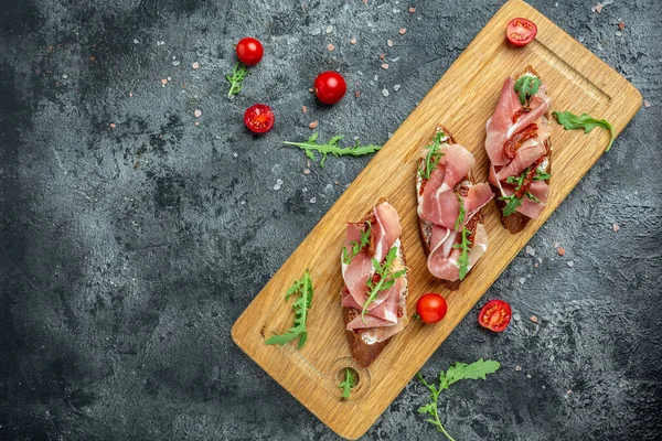 Kurutulmuş domates, prosciutto ve roka ile ahşap bir tahtanın üzerine konmuş bruschetta. Lezzetli kahvaltı, pankart, menü, metin için tarif yeri, üst görünüm,