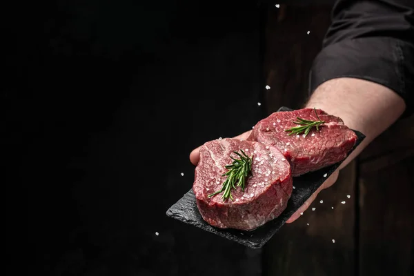 Chef salts steak in a freeze motion with rosemary and spices. Preparing fresh beef or pork on a dark background. Long banner format.