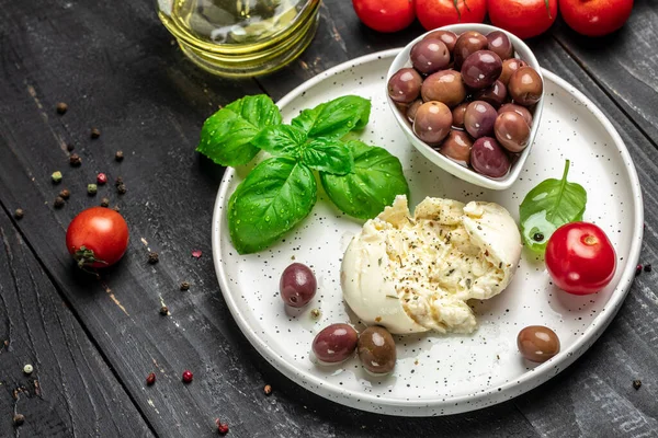 Kremalı İtalyan Burrata peyniri fesleğenli, zeytinyağlı ve beyaz tabakta vişneli domatesli koyu ahşap arka planda. afiş, menü, metin için tarif yeri, üst görünüm.