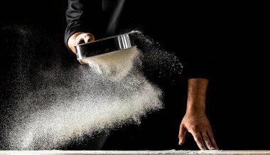 Şef eleğe un serpiyor, toz un havaya karışıyor. Un bulutunun dondurucu hareketinde un taşıyan şefin elleri.