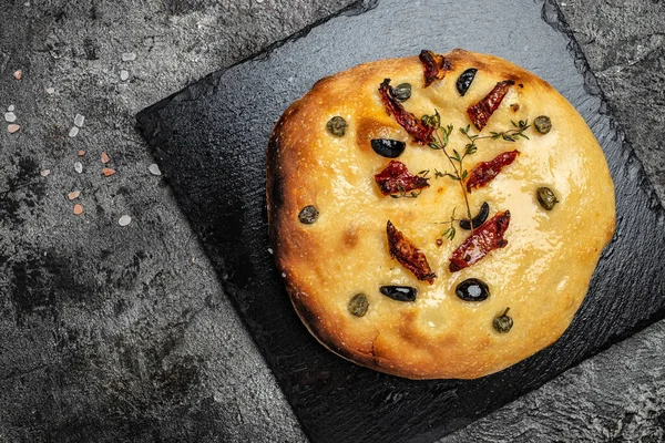 Foccacia, Italian bread, with olive oil, olives, sun-dried tomatoes, capers and thyme, Mediterranean bread on a dark background. place for text, top view.