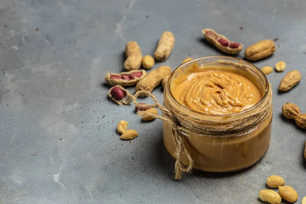 Peanut butter or paste in an open jar and peanuts in the peel scattered on a gray background, banner, menu, recipe place for text, top view.