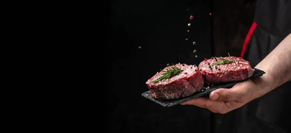 Chef salts steak in a freeze motion with rosemary and spices. Preparing fresh beef or pork on a dark background. Long banner format.
