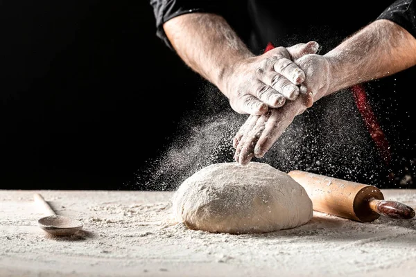 Beautiful and strong men\'s hands knead the dough from which they will then make bread, pasta or pizza. A cloud of flour flies around like dust. Food concept.
