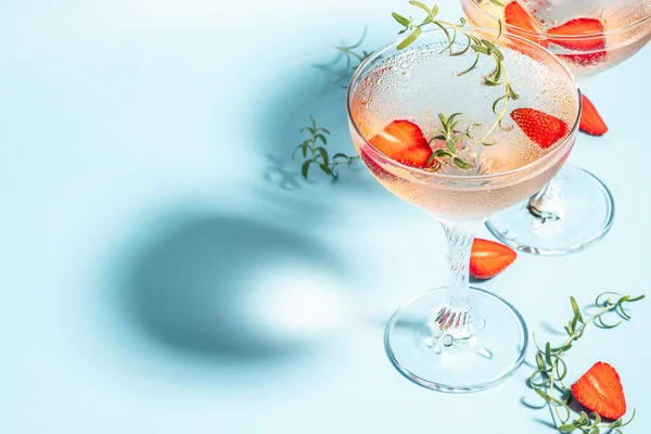 Glasses with cold pink champagne or punch with wine strawberries, ice and rosemary on blue background.