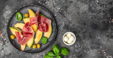 Prosciutto ve fesleğenli taze kavun. Antipasti, geleneksel İspanyolca ve İtalyan aperatifleri. Koyu taş masadaki üst manzara, uzun pankart biçimi. en üstteki görünüm.