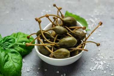 Kapari. Salamuraya yatırılmış ya da turşulu kapari meyvesi. Küçük bir kasede taze fesleğen yaprağı, gri arka planda. Uzun afiş biçimi. en üstteki görünüm.