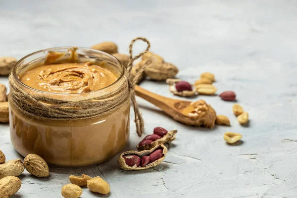 peanut paste in an open jar in spoon near creamy peanut butter peanuts scattered on a light background, place for text, top view.
