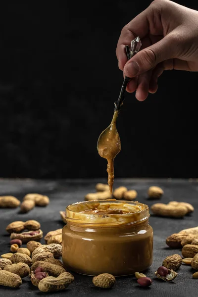 peanut paste in an open jar in spoon near creamy peanut butter peanuts scattered on a dark background, place for text, top view.