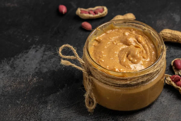 Peanut butter or paste in an open jar and peanuts in the peel scattered on a dark background, banner, menu, recipe place for text, top view.