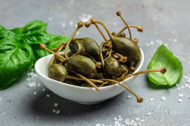 Kapari. Salamuraya yatırılmış ya da turşulu kapari meyvesi. Küçük bir kasede taze fesleğen yaprağı, gri arka planda. Uzun afiş biçimi. en üstteki görünüm.