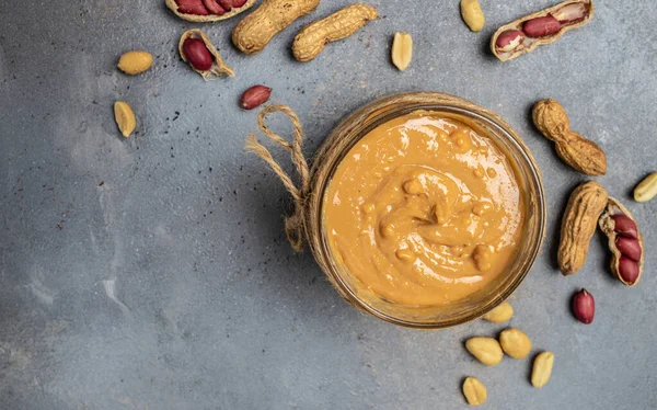 peanut paste in an open jar. creamy peanut butter peanuts scattered. Long banner format. top view.