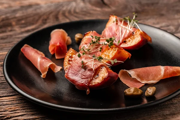 Peach slices wrapped in prosciutto decorated with with microgreens and capers. Fruit and Meat.