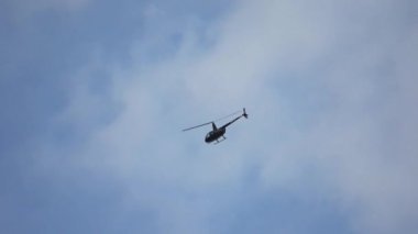 Küçük siyah bir helikopter mavi ve bulutlu bir gökyüzünde bir eğri üzerinde uçuyor..