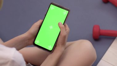 Çocuk jimnastik paspasında oturuyor ve yeşil ekranlı akıllı telefonu tutuyor.